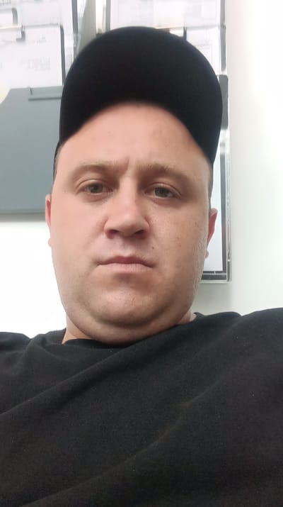 A man wearing a black t-shirt and a black baseball cap is shown in a close-up, head-and-shoulders shot. The man has fair skin and light brown hair. He is looking directly at the camera with a neutral expression. The background consists of a wall with some papers on it and a white wall on the right. The lighting is bright, suggesting an indoor setting, possibly an office or a residential room in Horný Hričov, Slovakia. The image is taken from a low angle.
