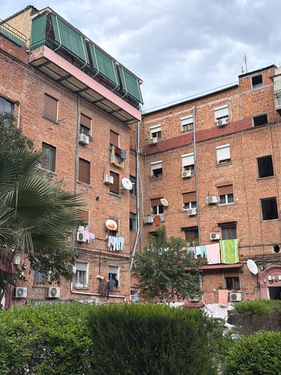 The image depicts a multi-story brick apartment building in Tirana, Albania. The building's exterior features numerous windows, many with closed shutters or blinds, and several air conditioning units mounted below them. Some windows have satellite dishes attached nearby. Clotheslines with laundry are strung between some windows. The building appears to be located in a residential area, with trees and greenery partially obscuring the view. The sky is overcast with clouds. The building's architecture is typical of residential structures in the city.