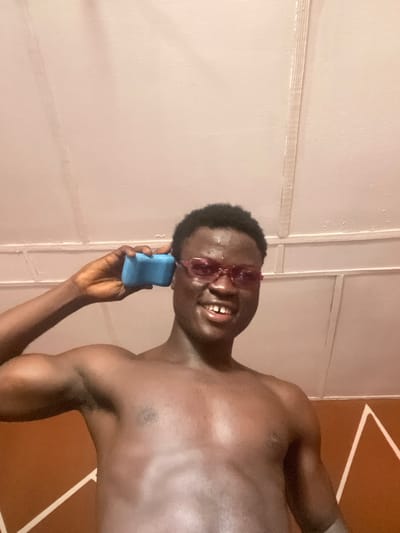 A male subject with dark skin, short dark hair, and pink-tinted glasses is depicted smiling directly at the viewer from a low-angle perspective. He has a visible dental gap between his upper front teeth and moisture on his forehead. The subject is shirtless, displaying his upper torso and arms. He holds a light blue, rectangular, porous object, resembling a sponge, against his left ear. The background consists of a light-colored paneled ceiling or upper wall with visible structural lines, and a section of a brown wall with a white geometric pattern is visible on the lower right. The scene is located indoors in Mosafejaw, Nigeria.