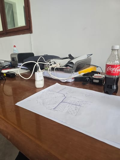 The scene depicts a cluttered office or workspace in Nyíregyháza, Hungary. A brown wooden desk is partially visible, covered with various objects. On the right, a clear plastic bottle of Coca-Cola, with a red label displaying "Coca-Cola" and "EREDETI ÍZ" in white text, is approximately one-third full. To its left, a black and red laser distance meter or measuring tape lies on the desk. In the foreground, a white sheet of paper displays handwritten blue ink drawings and numerical notations, including grid patterns, measurements like "50cm" and "B50 x 4.72", and the number "245".
Further left, a white "delight" branded USB wall charger is plugged into a white two-prong power adapter, which rests on the paper. A white USB cable extends from the charger towards the left. Behind these, a cluster of electronic devices and office supplies is visible, including a black power adapter, a black soft case or bag, and various papers and documents. A small, grey-colored electronic device with a display is positioned behind the black bag, and a red-capped bottle containing a light blue liquid (likely cleaning solution) is on its left.
The background consists of a plain white wall. In the upper left quadrant, a brown wooden-framed window with translucent or reflective glass is visible, suggesting an interior setting with natural light source. No individuals are present in the image.