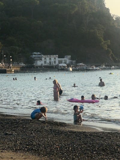 The image depicts a daytime coastal scene in Sestri Levante, Italy, specifically from the Baia delle Favole (Bay of Fables) looking towards the peninsula. The foreground features a dark pebble beach where two children are present. One child, wearing blue shorts, is squatting near the waterline, appearing to interact with the pebbles. Another smaller child, in a light-colored swimsuit, sits at the water's edge, holding a blue container.
The mid-ground shows the calm, light blue water of the bay, populated by approximately 15-20 individuals. Many are wading or swimming; a woman in a pink swimsuit stands upright in mid-thigh deep water, facing left, with another person partially obscured beside her. A pink inflatable raft is visible with at least one person on it and others in the water nearby. Yellow buoys are scattered across the water.
In the background, on the distant shoreline of the peninsula, a prominent white building with arched windows on its upper floor is visible, indicative of a club or establishment like the Circolo Velico Luigi Sirena. To the left of this building, a quay or pier with people and lampposts extends into the water. Numerous small boats, including motorboats, are moored in the bay adjacent to the distant shore. A dense, dark green, tree-covered hillside rises steeply behind the distant buildings and shoreline. The scene is illuminated by natural light, suggesting late afternoon or early morning, with the distant hillside in partial shadow.