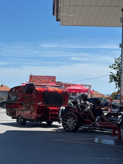 In the city of Byala, Bulgaria, on a bright, sunny day, a custom red and black flame-patterned trailer is parked next to a trike with two occupants wearing helmets. The trailer has a small window on the side and a storage compartment. A red awning with "Coca-Cola" branding is visible behind the trailer. To the left of the trailer, a dark-colored car is parked. Further in the background, several buildings with red tile roofs are visible. The scene appears to be near a gas station, as indicated by the overhead structure and a fuel pump in the foreground. A sign above the trailer reads "БЪРЗО И ВКУСНО" (Fast and Delicious) and lists food items such as "СУПА" (Soup), "СКАРА" (Grill), "ГОТВЕНО" (Cooked), and "ПИЦИ" (Pizzas). The sky is clear blue with a few wispy clouds.