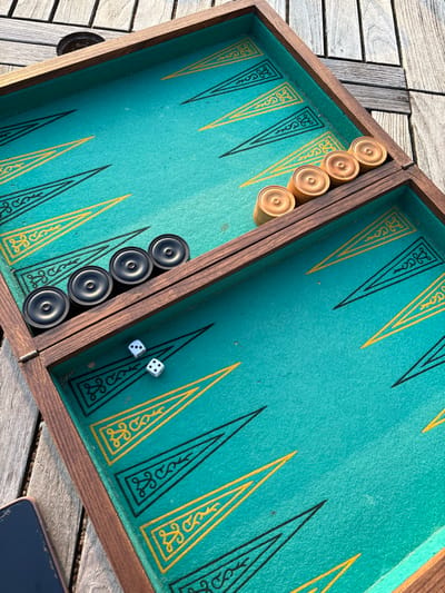 An elevated, downward-angled photograph captures an open backgammon board resting on weathered wooden decking outdoors in Bû, France. The board features a dark brown wooden frame surrounding a teal felt playing surface. The points are delineated by alternating black and yellow triangles, with the yellow points containing intricate scrollwork designs. Six black circular checkers are stacked along the left side of the central bar, closer to the bottom-left corner of the board's interior. Four light-colored circular checkers are stacked along the right side of the central bar, closer to the top-right corner of the board's interior. Two white dice rest on the teal felt in the bottom-left quadrant of the board's playing surface, with one die displaying a single pip and the other displaying two pips. The surrounding environment consists of visible wooden planks, indicating an outdoor setting, illuminated by natural daylight. No individuals are present in the image.