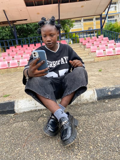 A young person with dark skin and braided hair sits on the ground in front of bleachers. They are wearing a black long-sleeved shirt with white lettering and dark wash wide-leg jeans. The person is holding a smartphone in their left hand and looking at the screen. They are also wearing black platform loafers and grey socks. The background consists of pink bleachers and a building with yellow walls and windows. The ground is paved with asphalt and gravel, and there is a curb with black and white markings. The scene appears to be outdoors, possibly at a sports stadium or school in Zawan, Nigeria.