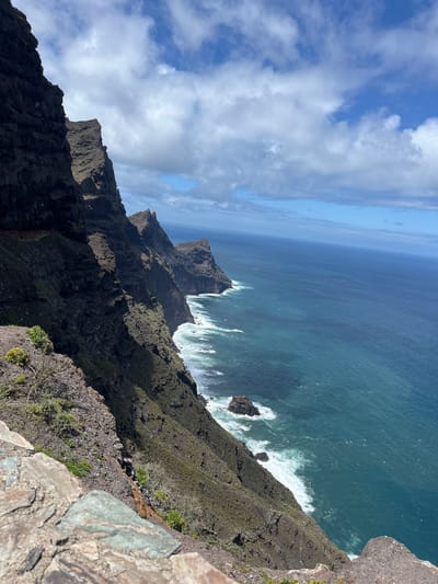The image captures a dramatic view from a cliff overlooking the coastline of Agaete, Spain. The vantage point is high, providing a steep downward angle onto the dark, rugged cliffs that plunge into the turquoise ocean. White waves crash against the base of the cliffs and rocky outcrops. Above, a blue sky is interspersed with patches of white clouds. The foreground features stone and rocky terrain with sparse vegetation. The photo emphasizes the ruggedness of the terrain, contrasting the rough cliffs with the sea and sky.
