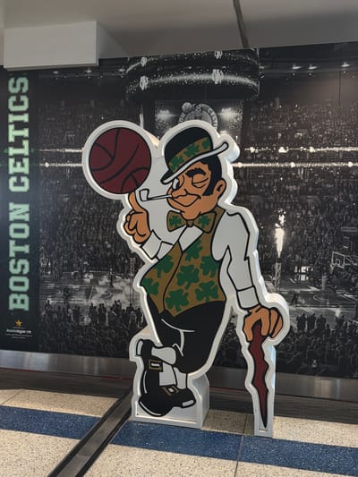 A life-size, cutout figure of a leprechaun, representing the Boston Celtics, stands in front of a large mural depicting a basketball game. The leprechaun is wearing a green vest with shamrock patterns, a white shirt, black pants, and a black hat. He is holding a pipe in his mouth and a cane in his right hand. The mural behind him shows a crowd of people in a stadium and a basketball court. The Boston Logan International Airport is identified on a sign at the bottom left of the mural. The scene appears to be an indoor public space, possibly an airport terminal.
