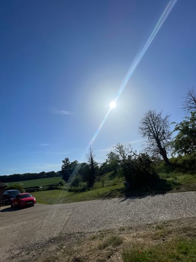 The image depicts a rural scene in Ashford, United Kingdom, on a sunny day with clear blue skies. A bright sun with a noticeable lens flare dominates the upper left portion of the frame. A narrow paved road runs along the right side of the image, bordered by grassy verges and hedgerows. Parked on the road at the bottom of the image are two cars, one red and one dark-colored. Beyond the road, a field of green grass extends towards a treeline. Several bare trees are visible, silhouetted against the bright sky. The image is rotated, making the sky appear as the left side of the frame.