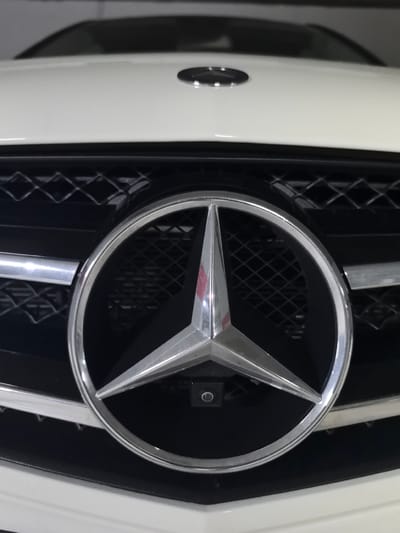 The image shows a close-up of the front grill and hood of a white Mercedes-Benz automobile. The Mercedes-Benz emblem is prominently centered in the frame. The background consists of the car's grill and the hood, with the top of the windshield visible. The image was likely taken in a parking structure given the muted light and general darkness in the background. The location in Rubí, Spain cannot be determined from the information provided in the image. There are no people visible in the image.