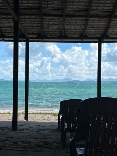 The image shows a view of the ocean from a covered outdoor seating area. In the foreground, two dark plastic chairs are partially visible, facing away from the camera. The floor of the seating area appears to be sand. Beyond the chairs, a sandy beach leads to the turquoise ocean. Gentle waves are breaking on the shore. In the distance, a line of hazy mountains or islands forms the horizon under a bright blue sky with scattered white clouds. The covered area has a tiled roof supported by dark wooden posts. The overall scene suggests a tropical beach location. The city of Ban Khlong Hia, Thailand is a coastal area known for its beaches and ocean views, making this a plausible scene for that location.
