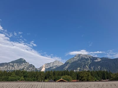 The image captures a panoramic view of mountains in the distance, with a clear blue sky dotted with wispy white clouds overhead. In the foreground, a tiled roof of a building is visible. A dense forest of green trees separates the roof from the majestic mountains. The mountains themselves are rugged and appear to be composed of rock and vegetation. One of the mountains has a prominent section of exposed light-colored rock, suggesting a quarry or a landslide. The overall scene is serene and depicts a natural landscape. The location is Münster, Austria.