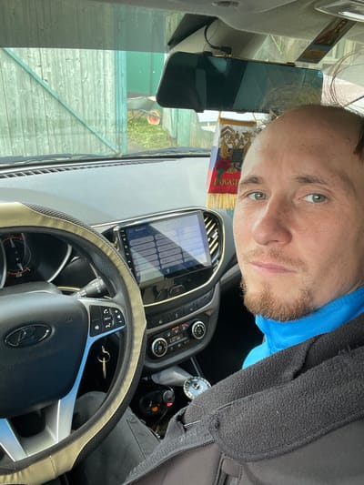 A man with light brown hair and a light beard is sitting in the driver's seat of a car, looking at the camera. He is wearing a blue turtleneck and a black zippered jacket. The car's dashboard is visible, including the steering wheel, a car stereo with a touchscreen display, and climate controls. A flag with the Russian coat of arms is hanging from the rearview mirror. Outside the car, a green wooden fence and a wheelbarrow are visible. The image was taken in Nizhnii Bugrysh, Russia.