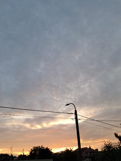 A silhouetted lamppost stands to the right of the frame against a sky filled with clouds, tinged with the warm hues of sunset. Electrical wires stretch horizontally across the sky, passing in front of the lamppost and extending towards the left edge of the image. Below the wires, the outlines of residential buildings and trees are visible against the lighter sky. The overall scene suggests a view of a residential area in Mława, Poland, during twilight.