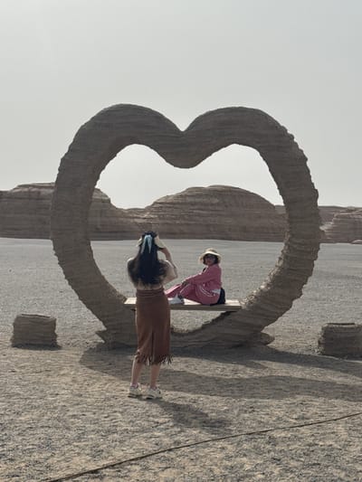 The photo shows two people interacting with a large stone heart-shaped sculpture in Dunhuang City, China. One person, dressed in a brown skirt and blue hair wrap, faces the heart and appears to be taking a photograph. The other person, wearing a pink tracksuit and straw hat, is situated within the heart itself, possibly posing. The sculpture is made of rough-hewn stone and is set against a backdrop of rocky formations and a pale sky. The ground around the sculpture is also made of stone.
