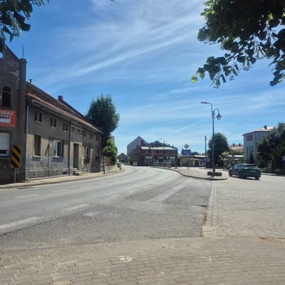 The image depicts a street scene in Bralin, Poland, on a sunny day with a partly cloudy blue sky. In the foreground, a paved sidewalk lines a street that curves to the right. A green car is parked on the right side of the street, facing away from the viewer. On the left side of the street, a gray building with a red-tiled roof and arched windows stands. Further down the street, other buildings and trees are visible. Traffic signals and streetlights are present. The overall scene is a typical view of a town or village street.