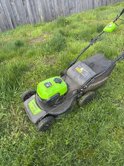 An overhead, slightly tilted shot captures a Greenworks lawnmower resting on overgrown grass next to a rustic wooden fence in what appears to be a yard or garden in Kuda, Russia. The scene is outdoors under an overcast sky, indicated by the soft, diffused lighting and lack of harsh shadows.
The primary subject is a black and bright green battery-powered Greenworks lawnmower. Its main body is black with a large lime-green battery cover or engine housing clearly visible towards the front. The grass is long and uneven, with scattered patches of small yellow wildflowers, possibly dandelions, particularly visible in the foreground and near the fence. Some areas of the grass appear slightly matted down, suggesting movement of the mower, or perhaps just uneven growth. The mower's wheels are dark and show signs of use, with some dirt and grass clinging to them.
Visible text on the mower includes "Greenworks" logos on the grass bag, the front deck, and the battery housing. The battery housing also displays "40V" and "Brushless," indicating its power source and motor type. Several warning symbols are present, including those for ear protection, foot protection, keeping bystanders away, and a recycling symbol. The noise level "96 dB" is also marked. The cutting width "46 cm" is visible on the front of the mower deck.
No people are visible in the image. The wooden fence, composed of weathered gray planks, runs along the left side of the frame, providing a boundary to the grassy area. The overall impression is one of a typical yard chore awaiting completion or having just started.