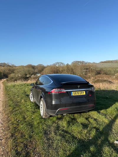A black Tesla Model X, bearing UK registration plate GE19 ERY, is stationary on a grassy verge adjacent to a dirt track under a clear blue sky. The vehicle is positioned facing away from the camera, at a slight angle showing its rear-left quarter. A red "Tdi" badge is affixed to the lower right of the boot lid. The car's lower body shows evidence of light dirt accumulation.
The immediate ground consists of vibrant green grass, interspersed with some dry patches, and a prominent brown dirt track occupying the left foreground and extending into the middle distance. Sharp shadows from an unseen object (likely the photographer) are cast across the grass towards the lower right.
The background features a dense line of trees and shrubs, primarily deciduous and bare, indicating a late autumn or winter season. Through this foliage, a light-colored building with a dark roof is partially visible on the right. Another lighter, smaller structure is also discernible further left within the tree line. The sky is a uniform, cloudless light blue, with a single faint aircraft contrail visible in the upper right quadrant.
The scene's characteristics, including the unpaved track, grassy verges, and wooded backdrop with distant structures, align with the environment of **Lordswood Greenway** in Southampton, United Kingdom. No people or active interactions are present.