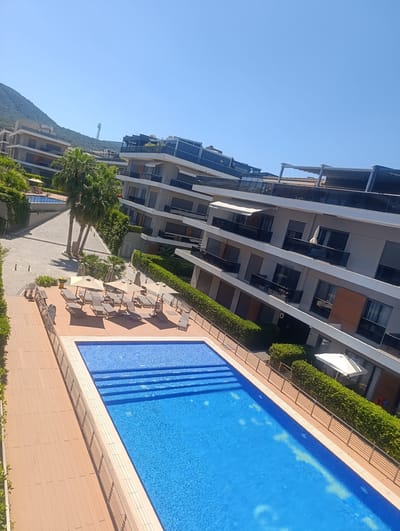 This media file shows a serene outdoor scene on a bright, sunny day in Urla, Turkey. The primary subjects are a swimming pool and a modern apartment complex. The pool, with its clear blue water and tiled steps, is the central focus, bordered by a wooden deck and a metal railing. Several lounge chairs and umbrellas are arranged on the deck, suggesting a relaxing atmosphere.

Behind the pool, lush green hedges surround the apartment buildings. The architecture is contemporary, with multiple stories, balconies, and large windows. Palm trees are interspersed among the buildings and greenery, adding to the tropical ambiance. In the background, a mountain covered in vegetation rises against a clear blue sky, providing a scenic backdrop. The time of day appears to be late morning or early afternoon, judging by the bright sunlight and the long shadows.

No people are visible in the media, emphasizing the tranquility of the setting. The overall mood is one of calm and leisure, suitable for a holiday destination. The presence of the apartment complex suggests a residential or resort area. The weather is clearly fair and warm, ideal for swimming and outdoor activities. There is a visible letter "D" on one of the building entrances.