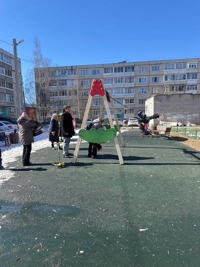 The image depicts an outdoor scene in Dobryanka, Russia, seemingly a playground or recreational area near an apartment building. Several people are present, including adults and children. A swing set, with a red and green top, is visible in the foreground, and people appear to be interacting with it. A stroller is parked nearby. The environment includes buildings, trees without leaves, and a paved surface. The weather is clear and sunny, casting shadows. The apartment building is characterized by its repetitive window patterns and a grayish facade.
