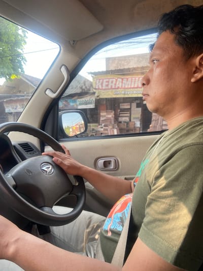 A man with dark hair is driving a car on a sunny day. He is wearing a green t-shirt and khaki pants, and a seatbelt is visible. The car's interior is visible, including the steering wheel, dashboard, and a side mirror. Through the side window, a street scene in Bangsal, Indonesia, can be seen, featuring buildings, trees, and signage that reads "KERAMIK" and "Jln. Raya Ban."