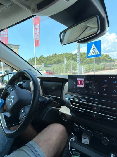 This media file captures an interior view of a car, likely taken from the driver's perspective while parked or driving in Mrčevac, Montenegro. The scene is set on a sunny day, suggested by the bright sky and clear visibility of the outdoor surroundings. Through the windshield, we see trees and buildings, hinting at an urban or semi-urban environment.
The focal point inside the car is the dashboard, dominated by a large touchscreen infotainment system displaying a radio interface with the station "PLAY Radio - Tivat / Herceg Novi - 104,6MHz". The screen also shows a temperature reading of 37°C and the car's air conditioning is set to AUTO. Beside the screen, various controls for climate and other functions are visible, including knobs and buttons.
The steering wheel, with the Citroën logo, is prominently featured. A portion of the driver's leg, clad in denim shorts, is visible on the right side of the frame, indicating the presence of at least one person in the vehicle.
Outside the car, visible through the windshield and side window, are road signs, including a blue pedestrian crossing sign. A large red banner with white lettering that appears to read "IDEA" is also visible, suggesting the car is near a commercial establishment, possibly a supermarket. The overall impression is a snapshot of a typical day spent traveling or stopped in a populated area, with clear weather conditions.