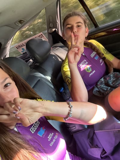 Two young women are sitting in the back seat of a car, likely in the city of Pirdop, Bulgaria, as indicated by a sticker on the rear window. The woman on the right is wearing a purple and yellow cycling jersey and is making a "shush" gesture with her fingers to her lips. The woman on the left is partially visible and appears to be covering her mouth with her hand, also wearing a similar cycling jersey. The interior of the car is visible, with black leather seats and the interior roof lining. Outside the car window, trees and sunlight are visible. The atmosphere suggests a candid moment, possibly during a break from a cycling event.