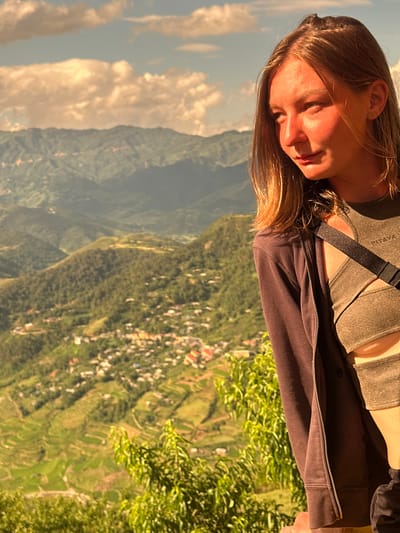 An outdoor, eye-level shot captures a young adult female positioned in the right foreground, looking downwards and to her left, with her profile partially illuminated by strong golden light. She has light brown hair and fair skin, with a warm glow on her right cheek and chin. She is wearing an olive-green textured crop top with cut-outs and a criss-cross design, featuring a faint "PITAYA" logo on the upper chest. A dark, wide strap crosses her chest, and she wears a dark brown/charcoal zippered hooded jacket.
The background reveals a vast mountainous landscape, characteristic of Tu Le, Vietnam. Multiple layers of green, forested mountains extend into the distance under a sky with scattered, warm-toned clouds. A village or town, consisting of numerous buildings with light-colored or red roofs, is visible on the lower slopes and in the valley. Distinct green rice terraces are identifiable on the lower mountain slopes, particularly towards the bottom left and center of the frame. Lush green foliage, including leafy branches, partially obscures the immediate foreground on the right. The scene is illuminated by strong, directional sunlight, suggesting late afternoon or early morning.