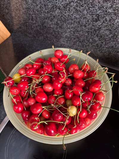 A bowl of fresh cherries sits on a dark, speckled countertop next to a wooden cutting board. The bowl is light green with a ribbed texture and is overflowing with bright red cherries, some with stems still attached. There is also a single yellow cherry in the bowl. The countertop appears to be granite or a similar material. The background is out of focus, showing a dark, textured surface that could be a wall or backsplash. The lighting is bright, casting shadows from the cherries. The image is taken from a high-angle perspective. The scene is located in Ilion, Greece.