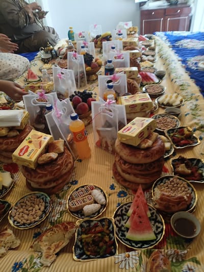 The image depicts a long, low table covered with a yellow and white patterned tablecloth, likely set for a meal. The table is laden with various food items, including bread, pastries, fruits, nuts, and what appear to be small dishes of cooked food. Several clear plastic bags with pink ribbons are distributed among the food, containing more pastries. In the background, a person in traditional clothing is visible, seated on the floor, and to the right, a blue patterned blanket is spread out. The overall impression is of a communal gathering or celebration. The scene is likely located in Osh, Kyrgyzstan, given the context.