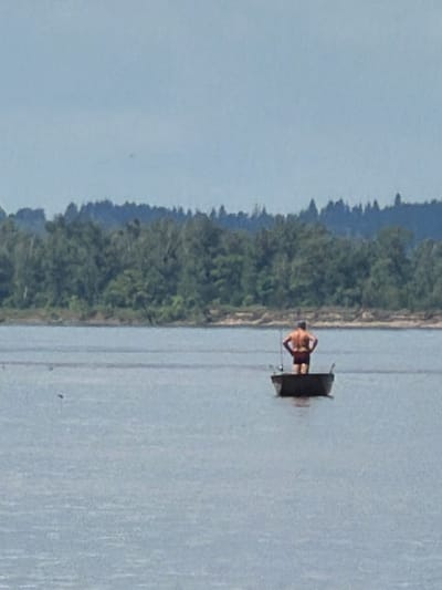 A single, shirtless male stands in a small, dark boat on a calm body of water. He is wearing dark shorts and has his hands on his hips. The boat is positioned towards the center of the frame, with a dense line of trees forming a dark silhouette across the horizon in the background. The sky is overcast with a pale blue hue. The water appears slightly rippled, and there are no other boats or people visible in the immediate vicinity. The overall scene suggests a quiet, possibly remote, natural setting.