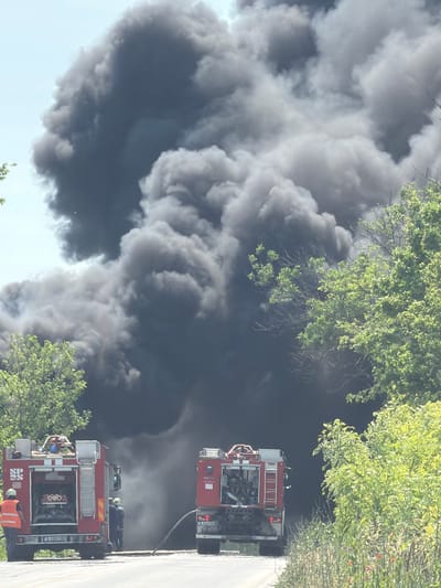 An emergency scene is depicted with a massive, dense plume of dark grey and black smoke dominating the central and upper portions of the frame against a light blue sky. The event is occurring in or near Topoli, Bulgaria. Two red fire trucks are positioned on a paved road in the foreground.

The fire truck on the left is oriented with its rear facing the viewer. It features a visible license plate reading "B 6082 HB" and a blue emergency light on its roof rack. Its rear compartment is open, revealing internal equipment. One individual wearing a high-visibility orange vest and a helmet stands to the left of this truck, facing its rear. Another individual in dark attire is partially visible near the truck's rear.

A second red fire truck is positioned on the right side of the road, further back and partially obscured by the smoke. A portion of its license plate, "D 0364 BA", is visible. A dark hose extends from this truck towards the area of heavy smoke. At least two individuals in dark uniforms or gear are partially visible near the rear of the right truck, appearing to handle equipment or hoses.

The paved road occupies the lower section of the image. Lush green trees and vegetation line both sides of the road, with particularly dense foliage on the right. The smoke appears to emanate from an unseen source beyond the immediate road area, rising significantly into the sky.