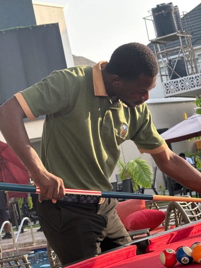 A man in Rubuchi, Nigeria, dressed in an olive green collared short-sleeved shirt with tan trim and black pants, is holding a pool cue. He is positioned next to a red pool table with several balls racked at the end. The background includes buildings, vegetation, and patio furniture. The setting appears to be outdoors.
