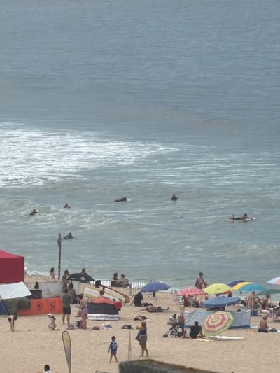 This is an aerial, high-angle, full shot of a crowded beach in Espinho, Portugal, during the day. The sandy beach is populated with beachgoers, tents, umbrellas, and beach equipment. The ocean in the background features numerous surfers and people swimming or wading in the water. The scene captures a typical summer day at a popular coastal location, with activities ranging from sunbathing to water sports.