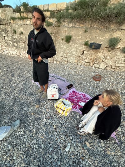 The image presents an overhead view of two individuals on a pebble beach or rocky coastline in Antibes, France, likely during the late afternoon or early evening. The ground is covered extensively with small, grey, and brown pebbles, and a low, weathered stone wall or cliff with sparse green vegetation borders the left side of the frame.

In the foreground, a man with dark hair and a beard stands, looking towards the camera. He is wearing a black zip-up jacket with some faint white embroidery (possibly "Sensation 72" or "22") and dark shorts, paired with light-colored, casual shoes. To his right, a purple and white striped blanket is spread on the pebbles, holding a white shopping bag clearly marked "ASIAKEO.com", a black flip-flop, and a small black device with "dbirmy" visible on it.

Further back and to the right, a woman with blonde hair tied back is crouched or sitting on a pink and purple patterned towel or blanket. She is dressed in a black jacket over a white long-sleeved top and white trousers, with patterned sneakers. She appears to be looking downwards, engaged with something in her lap or hands. Nearby, a brown woven object, possibly a small basket, and a yellow and white striped bag with "PAI. NIE" printed on it are visible. Another pair of feet, wearing light footwear, are seen in the bottom right corner of the image, suggesting the presence of a third person.

The overall atmosphere is casual and relaxed, indicative of people unwinding by the sea. The soft lighting suggests a pleasant, calm day without harsh sunlight.