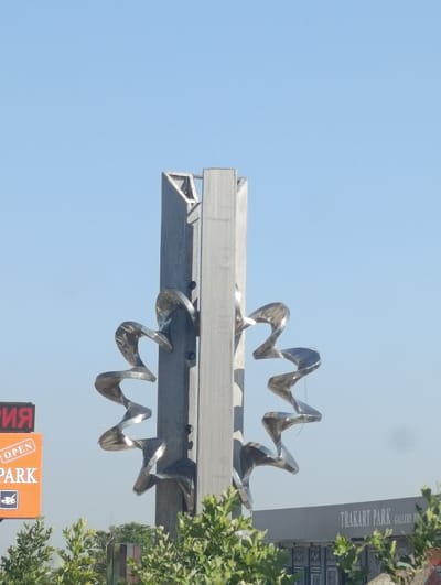 In the city of Krumovo, Bulgaria, a tall, metallic sculpture stands prominently against a clear blue sky. The sculpture is composed of a central, rectangular column with triangular elements at the top. Attached to the column, on both sides, are eight spiraling, metallic appendages that resemble abstract petals or wings. These appendages are attached at intervals, forming a circular pattern around the central structure.
To the left of the sculpture, a bright orange sign with white text reads "OPEN PARK." Above this sign, a portion of another sign is visible with red lettering that appears to read "РИЯ," suggesting a name or title in Cyrillic. In the foreground, lush green foliage partially obscures the base of the sculpture and the signs.
Further to the right and slightly behind the sculpture, a low, grey building is visible. White text on this building reads "TRAKART PARK GALLERY H." The building's facade features dark, patterned elements, possibly doors or decorative panels. The overall scene is an outdoor setting, likely a public space or an area associated with the "TRAKART PARK." The lighting suggests it is daytime, with no visible people or animals.