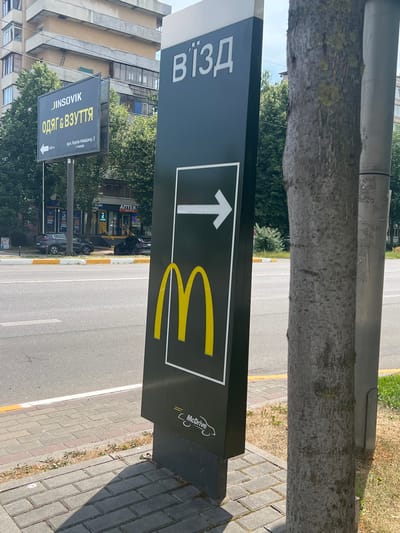 The image captures a street scene in Bucha, Ukraine. A tall, dark sign with white lettering indicating "В'їЗД" (Entrance) is prominently displayed, featuring a white arrow pointing to the right and a large yellow McDonald's logo. To the left of this sign, a separate billboard advertises "JINSOVIK - ОДЯГ & ВЗУТТЯ" (Clothing & Footwear) with an arrow pointing left and an address. In the background, buildings and trees line the street, and several cars are parked. The foreground shows a paved sidewalk and the road. The scene appears to be a sunny day.