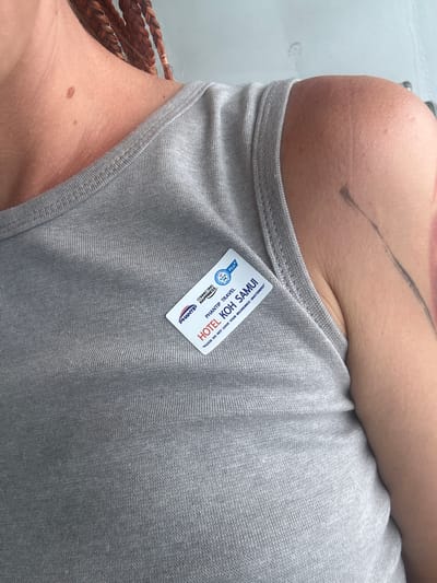 A close-up, upper-body shot captures a person wearing a gray tank top. A white name tag with blue and red lettering is affixed to the left side of the tank top. The name tag reads "PHANTIP TRAVEL HOTEL KOH SAMUI" and includes a small logo and the text "PLEASE DO NOT LEAVE YOUR BELONGINGS UNATTENDED". The person's skin is visible on their neck, shoulder, and arm, revealing several moles and a faint tattoo. The background is a light, neutral color, suggesting an indoor or overcast outdoor setting. The overall scene suggests the person may be a guest or employee of the hotel. The city of Don Sak, Thailand, is approximately 70 kilometers (about a 1-hour drive) from Koh Samui.