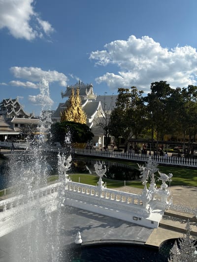 The image depicts a sunlit exterior view of Wat Rong Khun, commonly known as the White Temple, located in Ban Rong Khun, Chiang Rai Province, Thailand. In the foreground, a powerful fountain sprays white water upward from a dark pool, partially obscuring a white, intricately decorated bridge or walkway. This structure features numerous balusters and ornate mythological creature sculptures, appearing to be white Nagas or similar figures, on its railings.
In the midground, a serene, dark-water pond reflects the surrounding sky and structures. A pristine green lawn borders the pond, beyond which a white, ornate fence separates the lawn from a paved pathway. Numerous individuals, appearing as small figures due to distance, are visible walking along this pathway. Behind them, a line of dense green trees casts shadows, indicating direct sunlight from the upper right.
In the background, distinctive white temple buildings with multi-tiered roofs and elaborate gables are prominently featured. One central building displays a large, golden, flame-like sculptural element rising from its base. Other white buildings with similar architectural characteristics are visible further to the left. The sky is bright blue with scattered white cumulus clouds. The overall scene is well-lit, with strong highlights on the white structures and clear shadows from the trees and buildings.