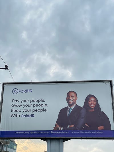 A billboard advertisement for PaidHR is displayed against a cloudy sky. The billboard features the PaidHR logo, a purple checkmark within a circle, and the company name. Below the logo, text reads: "Pay your people. Grow your people. Keep your people. With PaidHR." Two individuals, a man and a woman, are depicted smiling and looking towards the viewer. They are both dressed in formal attire. At the bottom of the billboard, contact information is provided: "Get Started Today," an email address "hello@paidhr.com," and a website address "www.paidhr.com." A slogan, "All-in-one HR software for growing teams," is also present. In the background, a glimpse of a city skyline with buildings is visible beneath the billboard, suggesting an urban environment, potentially Ojomu, Nigeria. Electrical wires and poles are visible above and to the left of the billboard.