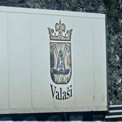The image displays a large, white rectangular panel, likely the side of a truck trailer, set against a rugged, rocky background with sparse green vegetation. The panel features a distinctive emblem and text.
The emblem, rendered in muted shades of brown, beige, and black, is composed of two main parts. On the left is an ornate, crown-like design with decorative curves and circular elements. To its right, a shield-shaped element contains an illustration of two crossed axes and a third tool resembling a pickaxe or mattock, all depicted with a rustic, textured appearance, including a prominent cross-hatch pattern on the central tool.
Immediately to the right of the emblem, the word "Valaši" is written in bold, black, stylized letters, with a caron (ˇ) above the first 'a'. The sign itself appears to be made of multiple horizontal sections. On the right side of the panel, dark, vertical structures are visible, consistent with the framework or other components of a vehicle.
The scene is illuminated by bright daylight, suggesting clear weather. There is no human activity or specific event taking place, solely the static view of the sign. The rocky terrain in the background indicates an outdoor setting, potentially a mountainous or hilly area, aligning with the context of Le Perthus, France.