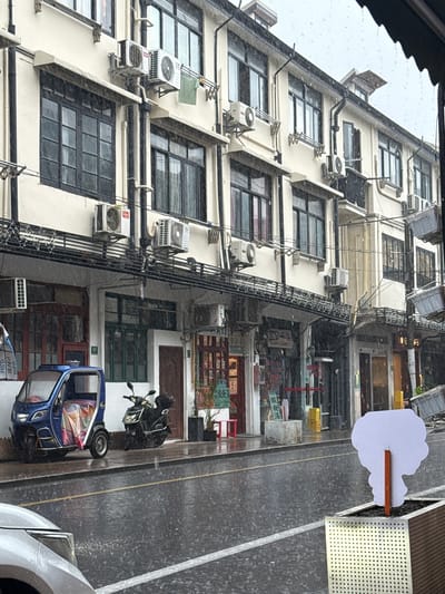 This image depicts a street scene in Xuhui District, China, during a rain shower. The focal point is a cream-colored, multi-story building with black trim and numerous windows. Several air conditioning units are mounted on the building's facade, and laundry is hung out to dry on a rack. Below the building, a row of shops or businesses are visible, some with their doors open. In the foreground, a blue three-wheeled vehicle is parked on the wet street, alongside a black motorcycle. The street is slick with rain, and visible raindrops are falling. A white car is partially visible in the lower left corner. A planter with a white, cloud-shaped sign on a stick is situated on the right side of the frame. The overall impression is a typical urban street environment experiencing inclement weather.