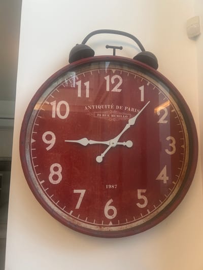 A large, round, red alarm clock is mounted on a white wall. The clock face has white numbers and hands. The top of the clock features two dark-colored bells and a handle. Text on the clock face reads "ANTIQUITE DE PARIS" and "28 RUE MURILLO" and "1987". The clock appears to be decorative and is not currently functioning. The environment is an indoor setting with plain white walls. There are no people or animals visible in the image. The scene does not provide any specific information about its location in the city of Hof HaCarmel Regional Council, Israel.