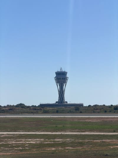 An air traffic control tower stands prominently against a clear blue sky. The tower, located in El Prat de Llobregat, Spain, features a distinctive X-shaped base supporting a cylindrical control cabin at the top. The surrounding environment consists of a grassy field in the foreground, with a paved runway or road visible beyond it. Further back, there is a line of trees and bushes, and behind that, a low-lying building structure. The overall scene depicts an airport environment under a bright, cloudless day.