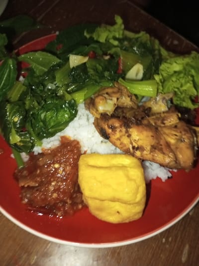 A close-up, elevated view captures a single serving of food presented on a circular red plate with a white outer rim. The plate rests on a dark, possibly wooden, surface under dim lighting. The meal consists of a central mound of white rice, flanked by several pieces of grilled or fried chicken with a dark golden-brown, crisp appearance. To the upper left and right, a substantial portion of vibrant green, stir-fried leafy vegetables, possibly bok choy, is arranged, featuring visible segments of what appear to be garlic and red chili. In the lower left quadrant of the plate, next to the rice, is a scoop of chunky, reddish-orange chili paste or sambal. Adjacent to the chili paste, in the lower right, sits a single, golden-yellow, rectangular piece of deep-fried tofu. No individuals or active interactions are visible in the scene. This prepared meal is observed in the city of Melakudikadu, India.