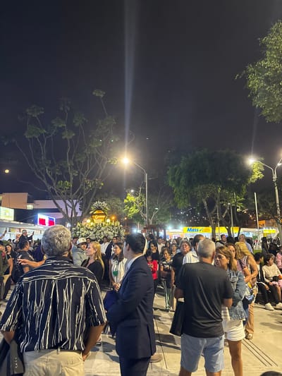 A crowd of people stands on a sidewalk in Ancón, Peru, at night. Streetlights illuminate the scene, casting light through trees and onto the faces of the gathered individuals. A floral arrangement is centered within the crowd's focus. Many are casually dressed in shorts and t-shirts. Buildings and businesses line the street. Some people appear to be looking toward the floral arrangement.