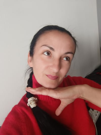 A medium shot captures a woman from the chest up, posing in front of a plain light gray wall. Her dark hair is pulled back and braided, with stray wisps framing her face. She wears a vibrant red turtleneck sweater. Her right hand rests beneath her chin, with her index finger extended and touching her cheek. Her eyes are looking upwards and to the right, suggesting contemplation or a moment of thought. The lighting appears natural, casting soft shadows. The image is a selfie, taken from a slightly low angle. The background is devoid of any discernible features, indicating an indoor setting. No information about the location in Czekanów, Poland, is available from the image itself.