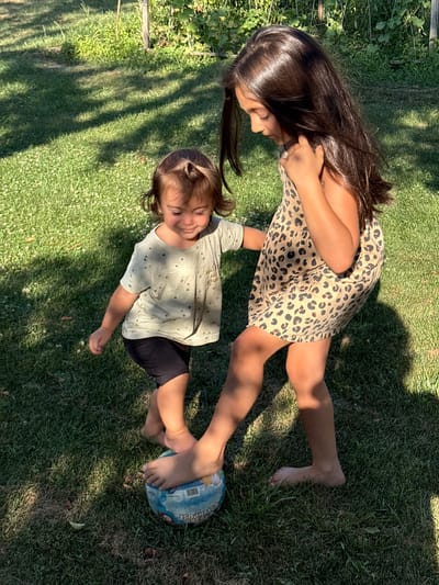 Two young girls are playing with a ball on a grassy lawn in a garden. The older girl, on the right, has long dark hair and is wearing a leopard-print dress. The younger girl, on the left, has dark hair and is wearing a light-colored t-shirt with small stars and black shorts. The older girl is standing with her foot on the ball, while the younger girl appears to be reaching for it. The background shows green grass, some foliage, and dappled sunlight and shadows. The image was taken in the city of Oreshak, Bulgaria.
