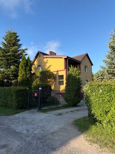 The image depicts a yellow house with a brown roof situated in Svetlogorsk, Russia. The house has several windows, some of which are visible from the exterior. The property is surrounded by lush green trees and hedges, creating a well-maintained garden setting. A gravel driveway leads up to the house, and a black metal gate with the number "13" displayed on it is visible at the entrance. The sky is a clear blue with a few wispy clouds. The lighting suggests it is daytime, with the sun casting shadows on the side of the house and the ground. The overall scene conveys a sense of suburban tranquility and domesticity.