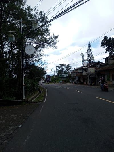 The image depicts a roadside scene in Phoxay, Laos, under an overcast daytime sky. The perspective is from a slightly elevated position, looking down a dark asphalt road that curves to the left.

On the immediate left, a dark, irregularly paved surface abuts a grass verge and dense, dark green foliage. A white-painted metal streetlamp post stands prominently, featuring two light green decorative lanterns and a circular white sign bearing dark Arabic/Jawi script. Tall pine trees are visible behind and above the streetlamp. Numerous black utility wires crisscross the upper portion of the frame.

The asphalt road features a continuous white line marking its left edge and a double solid yellow line visible further down the right lane. On the right side of the road, a red scooter with two helmeted riders (one with a white helmet, one with a pink/purple helmet) travels away from the viewer. Along the right side of the road, a series of single- and two-story buildings, appearing weathered with varied facades and roof materials including corrugated metal, line the street. Another white streetlamp, similar in design to the one on the left, is positioned among these buildings, alongside a tall, slender conifer tree and a small white satellite dish on a rooftop.

In the mid-distance, a large vertical billboard with a dark-toned advertisement is visible. Beyond the immediate buildings and trees, a distant range of hills or mountains is discernible beneath the cloudy sky.