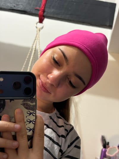 A woman with fair skin and dark eyebrows is taking a selfie. She is wearing a pink beanie and a striped shirt. She is holding a smartphone with a dark blue frame and a patterned case. The background includes a white wall, a dark window frame with a red string, and a white rope. There's an object on the top right corner of the photo.