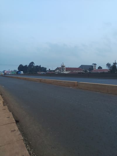 The image depicts a highway in Limuru Central ward, Kenya, under a cloudy sky. A concrete barrier separates the road from a paved shoulder. In the distance, several buildings are visible, including what appears to be a church with a steeple and other structures with red roofs. A few vehicles are present on the highway, including trucks and cars. Trees and utility poles line the background. The overall scene is that of a typical urban or suburban highway environment.